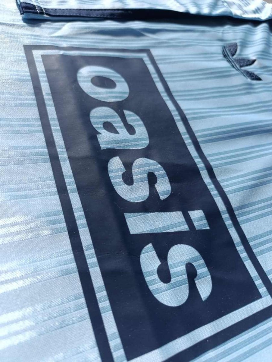 Aproximação do tecido celeste da camisa Oasis x Adidas com o trevo da Adidas Originals