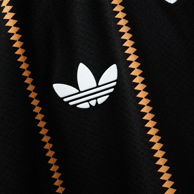 Detalhe do trevo branco sobre o tecido preto da camisa terceira da Alemanha 2026, entre as listras de losangos dourados