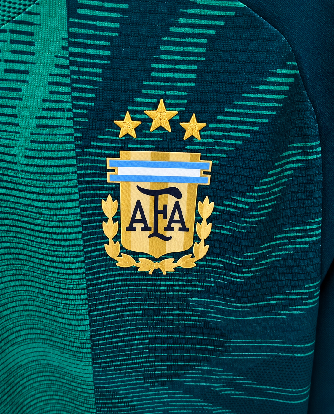 Detalle del escudo dorado de la AFA con tres estrellas sobre el tejido verde de la camiseta despedida de Messi
