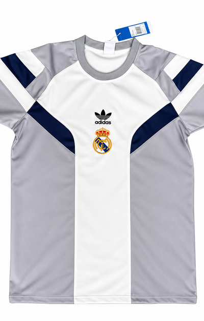 Camiseta retro del Real Madrid blanca con paneles grises y azul oscuro en hombros y costados, escudo dorado y trébol de la marca