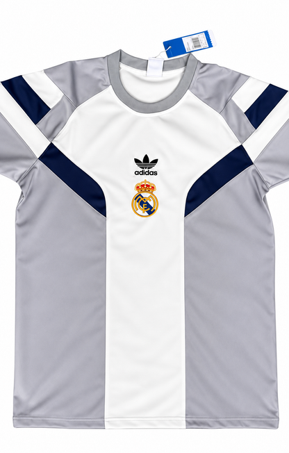 Camiseta retro del Real Madrid blanca con paneles grises y azul oscuro en hombros y costados, escudo dorado y trébol de la marca